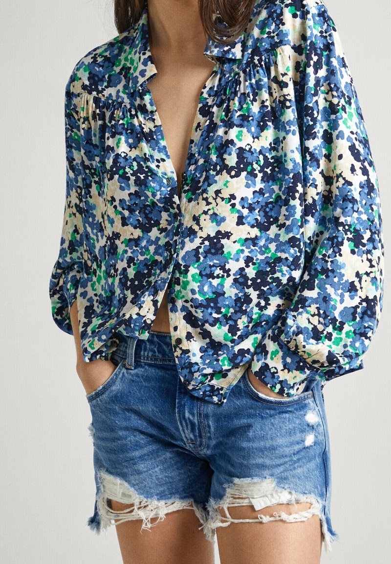 Femme portant un chemisier bleu à fleurs à manches longues rentré dans un short en jean bleu déchiré, les mains dans les poches.