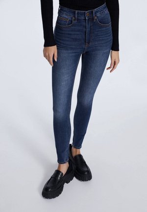 Personne portant un jean skinny bleu foncé, des mocassins noirs épais et un haut à manches longues noir, debout sur un fond clair uni.