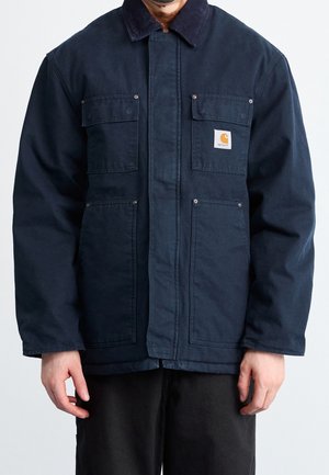 Mand iført en mørkeblå Carhartt jakke med fire forlommer og sorte bukser, stående mod en ensfarvet lys baggrund.