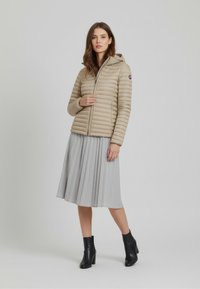 Beige gewatteerde jas met capuchon, ritssluiting aan de voorkant en verticale stiksels. Gecombineerd met een geplooide grijze rok en zwarte enkellaarsjes met hak.