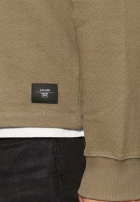 Sweatshirt vert olive avec des poignets et un ourlet côtelés, arborant une étiquette en tissu noir en bas à gauche, superposé sur une chemise blanche.