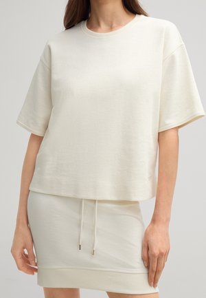 Vrouw draagt een loszittend crème kleurig top met korte mouwen en een bijpassende crème rok met trekkoord in de taille tegen een effen achtergrond.