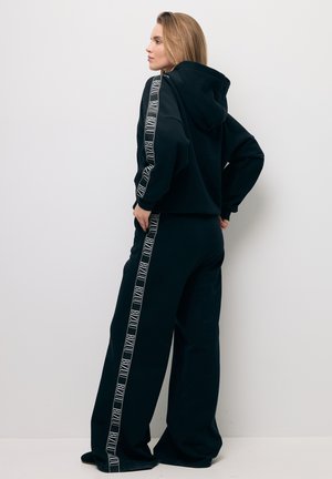 Kobieta w czarnej bluzie z kapturem i spodniach o szerokich nogawkach z białymi paskami z logo stoi bokiem przy białej ścianie.