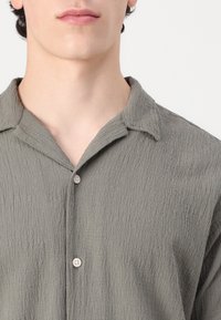 Camisa de botones de color gris claro con tela texturizada, que presenta un cuello puntiagudo y dos botones visibles en la tapeta.