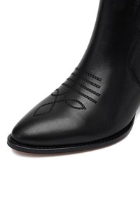 Mustad musta nahast pahkluu saabas, millel on terav varvas, dekoratiivsed tikandid ja peened reljeefsed mustrid varraste kohal. Sile tekstuur.