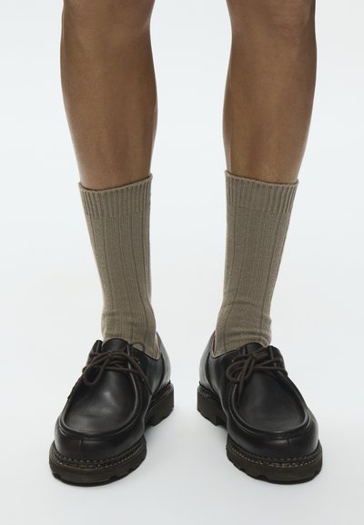 Jambes portant des chaussettes côtelées beiges à tige moyenne et des chaussures en cuir noires à lacets avec semelles épaisses sur fond blanc.