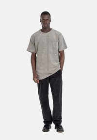 Graues Wildleder-T-Shirt mit Brusttasche, kombiniert mit schwarzen Cordhosen. Das Model trägt schwarze Schuhe. Einfaches, unverziertes Design.