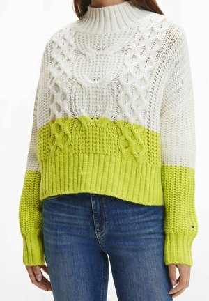 Pull tricoté blanc et vert citron avec motifs torsadés, porté avec un jean bleu, court à la taille et manches longues.