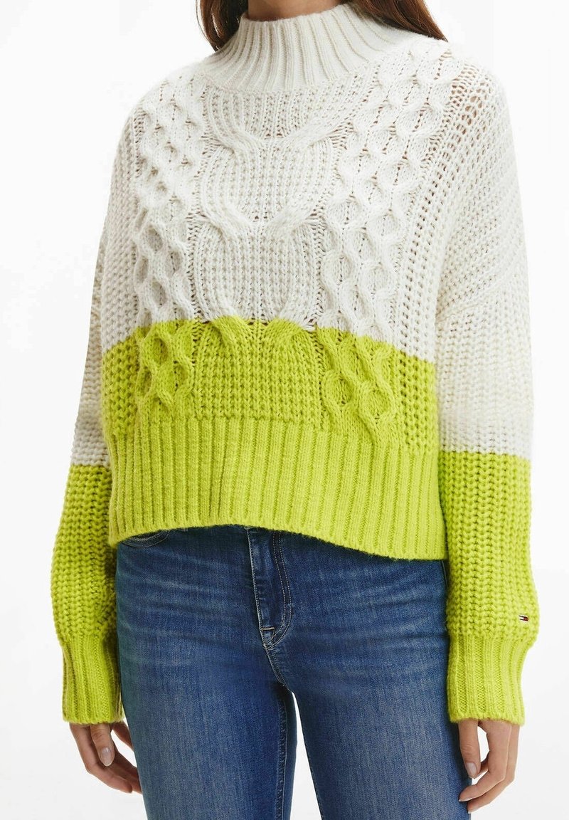Pull tricoté blanc et vert citron avec motifs torsadés, porté avec un jean bleu, court à la taille et manches longues.