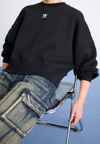 Svart sweatshirt med ribbad halsringning och muddade ärmar, med en liten vit Adidas-logotyp; bärs med slitna blå cargobyxor.