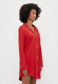 Cămașa de pijama roșie, fabricată dintr-un material moale, cu un guler și un față cu nasturi, are o buzunare cu broderie decorativă. Marginile sunt accentuate de cusături contrastante.