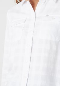 Vit skjorta med knappar, tillverkad av ett strukturerat tyg, med två bröstfickor och en liten logotyp på övre vänstra sidan. Lätt, andningsbart material.