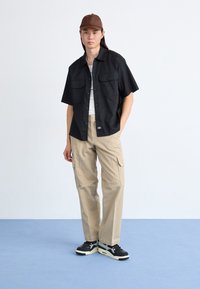 Sort kortærmet skjorte med to frontlommer, beige cargo bukser med sidelommer og sorte sneakers med lyse accenter. Brun kasket.