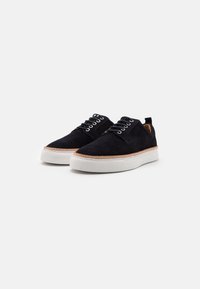 Sneaker basse in suede nero con suola in gomma bianca, allacciatura frontale e una piccola linguetta sul retro. Cuciture marroni accentuano la tomaia.