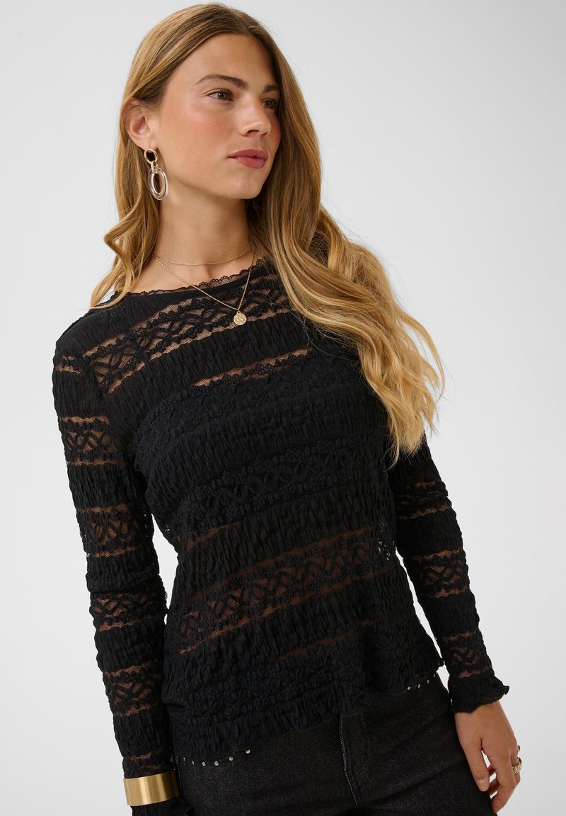 Femme aux longs cheveux blonds portant un top en dentelle noire à manches longues, des boucles d'oreilles anneaux dorées, un collier pendentif et un pantalon foncé sur un fond uni.