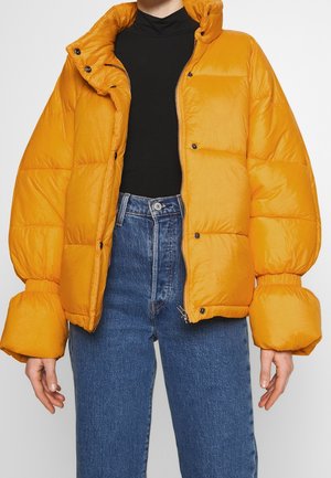 Puffet orange jakke med høj krave, lynlås lukning og store, samlede ærmer; båret over en sort top og blå jeans.