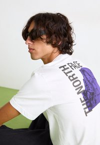 Hvid bomulds T-shirt med et lilla grafisk tryk af et bjerg og teksten "THE NORTH FACE" i fed sort på ryggen.