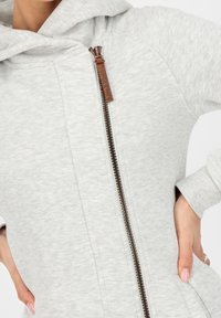 Vaaleanharmaa vetoketjullinen huppari, joka on valmistettu pehmeästä kankaasta. Siinä on etuvetoketju, ruskea yksityiskohta, huppu ja ribbireunat.