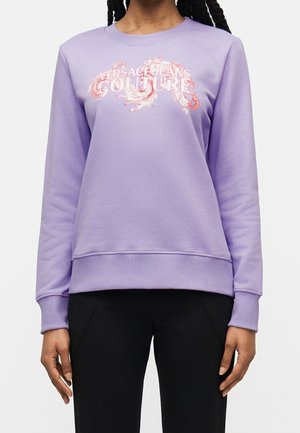 Personne portant un sweat-shirt lavande avec le texte "Versace Jeans Couture" et un motif décoratif, associé à un pantalon noir, debout devant un fond uni.