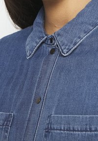 Jeansskjorta med spetsig krage, svarta tryckknappar och två bröstfickor. Mörkblå tyg med subtil struktur och sömnadsdetaljer.