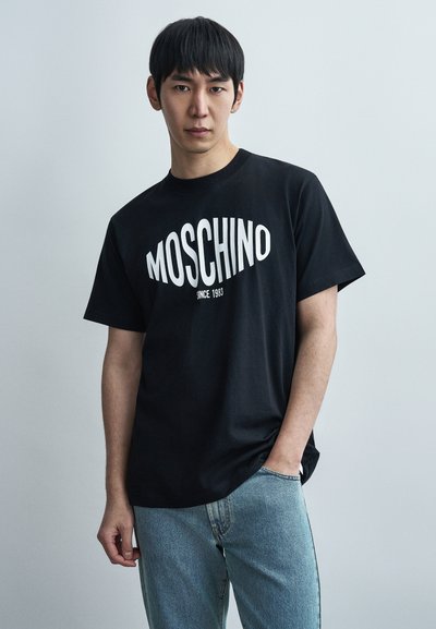 Μαύρο βαμβακερό t-shirt με έντονο λευκό λογότυπο "MOSCHINO" και κείμενο "SINCE 1983", συνδυασμένο με ανοιχτόχρωμο τζιν παντελόνι.
