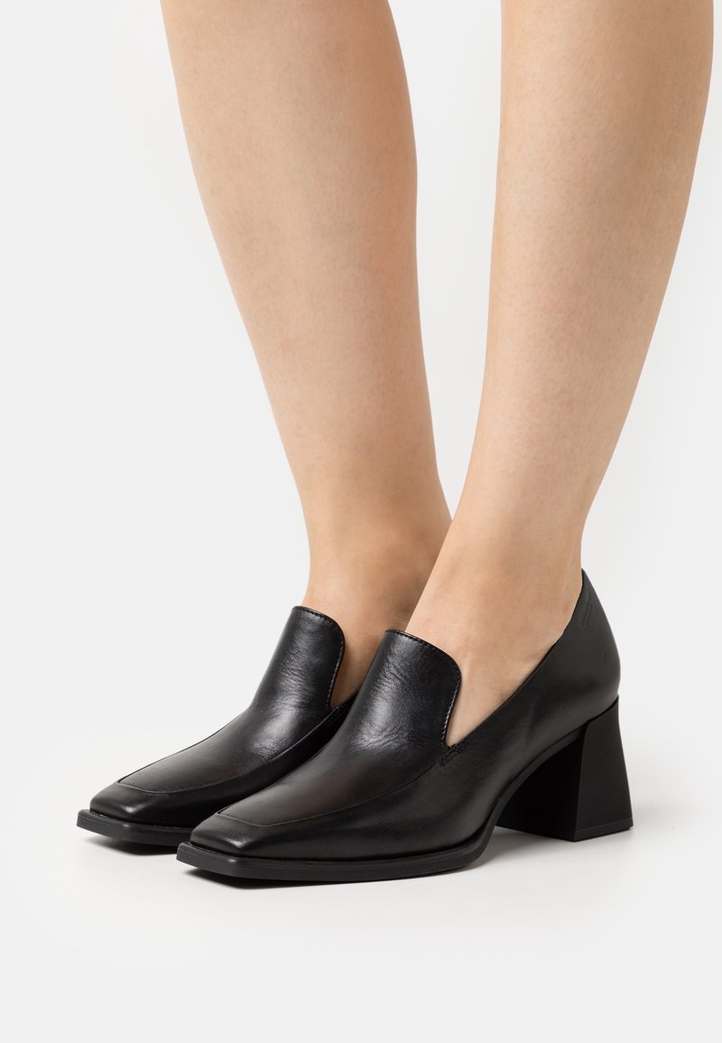 Chaussures en cuir noir à talon moyen, faciles à enfiler, portées sur jambes nues, avec des bouts carrés et un talon bloc, présentées sur un fond clair uni.