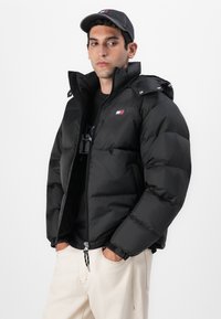 Svart boblekåpe med glidelås foran, hette og trykknapper. Har quilted detaljer og et lite logo-patch på brystet.