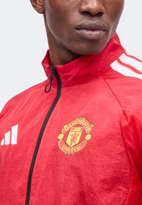 Άνδρας με κόκκινο μπουφάν προπόνησης Manchester United, με μαύρο φερμουάρ και λευκές ρίγες Adidas στον ώμο, στραμμένος ελαφρώς προς τα δεξιά.