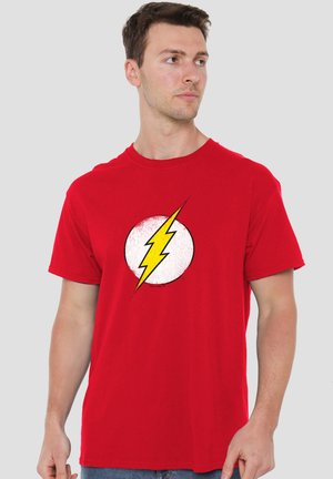 Mann trägt rotes T-Shirt mit weißem Kreis und gelbem Blitzsymbol, blickt seitlich vor schlichtem grauem Hintergrund.