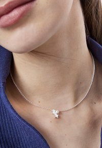 Srebrny naszyjnik z delikatnym łańcuszkiem, z małym charms w kształcie gwiazdki i perłowym akcentem. Noszony na tle strukturalnego granatowego swetra.