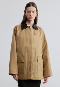 Μπεζ εξωτερικό μπουφάν με μαύρο γιακά, με κουμπιά τύπου snap και δύο μπροστινές τσέπες. Δομημένος, ελαφρώς φαρδύς σχεδιασμός με μακριά μανίκια.