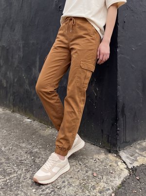 Personne portant un pantalon cargo marron et des baskets beiges, appuyée contre un mur texturé sombre sur un sol en béton brut.