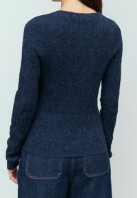 Pull texturé bleu marine à manches longues avec une coupe ajustée, des détails côtelés et un col rond, associé à un jean en denim bleu.