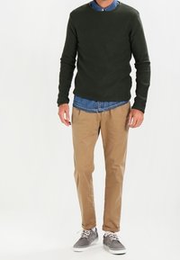Maglione a coste verde scuro indossato sopra una camicia di denim azzurro chiaro, abbinato a pantaloni chinos beige e sneakers grigie con suole e lacci bianchi.