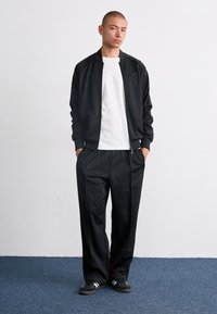 Training sportiv negru, compus dintr-o jachetă cu fermoar și pantaloni asortați dintr-un material neted. Sub jachetă se află un tricou alb, completat cu pantofi sport negri.