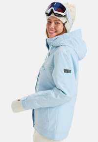 Helle blaue Skijacke mit Kapuze, ausgestattet mit einer Frontreißverschlusstasche, seitlichen Reißverschlüssen und einem kleinen Logopatch am Ärmel. Handschuhe und eine Skibrille sind sichtbar.