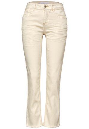 Nahaufnahme einer Person, die cremefarbene Jeans mit Gesäßtaschen und einem Etikett am Bund trägt, kombiniert mit einem goldenen Armband am rechten Handgelenk.