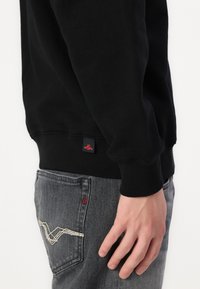 Sudadera negra con puños acanalados, combinada con unos vaqueros grises que tienen un patrón decorativo de puntadas blancas en el bolsillo trasero y un acento rojo.