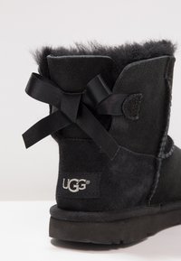 Svart mocka ankelstövel med en mjuk, foder av päls i överkant. Har en svart satinrosett och en UGG-logopatch på sidan. Gummisula.