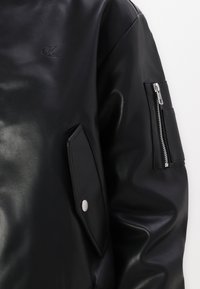 Giacca in pelle nera con una texture liscia, tasca con cerniera sulla manica e tasca con pattina, con un logo tondeggiante impresso sul davanti.