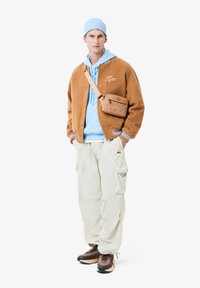 Giacca varsity in misto lana marrone, felpa con cappuccio azzurro chiaro, pantaloni cargo bianchi, scarpe da ginnastica marroni con motivo e borsetta a tracolla beige con grafica.