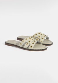 Lichtbeige instapsandalen met drie brede banden versierd met gouden studs en een lichtgeïsoleerde voetbed op een platte zool.