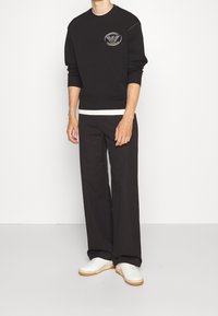 Felpa nera con collo rotondo e patch con logo, abbinata a pantaloni neri larghi e sneakers bianche con suola beige.