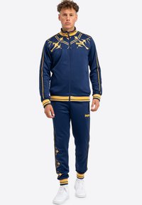 Survêtement marine avec accents jaunes ; veste zippée avec un design graphique sur la poitrine et des poignets rayés ; pantalons à taille élastique avec logo.