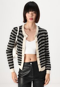 Zwart en crème gestreepte cardigan met een gebreide textuur, voorzien van een ronde hals, knoopsluiting en geribbelde randen. Gecombineerd met een wit topje.