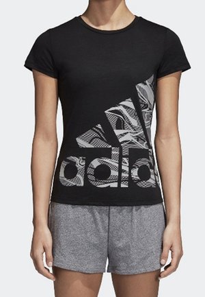 Frau trägt ein schwarzes, eng anliegendes T-Shirt mit großem weißem Adidas-Logo und graue Sportshorts, steht mit entspannten Armen an den Seiten.