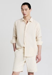 Giovane uomo con capelli slicked-back che indossa una camicia a maniche lunghe con bottoni di colore crema texturizzata e pantaloni corti abbinati, in piedi con una mano in tasca.