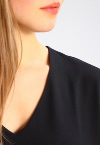 Musta, sileä kangas v-kaula-aukkoinen paita, jossa on hieno kiilto, rento kaula-aukko ja pehmeä rakenne. Hiukset valuuvat pehmeästi olkapäälle.