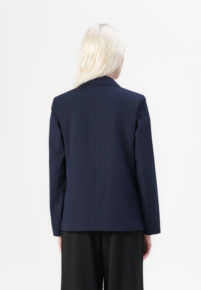 Blazer marine à coupe ajustée, avec revers crantés, fente unique au dos et épaules structurées. Texture de tissu tissé lisse.