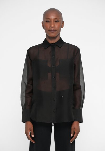 Femme avec la tête rasée portant une chemise noire transparente à manches longues boutonnée sur un crop top noir, se tenant devant un fond blanc.